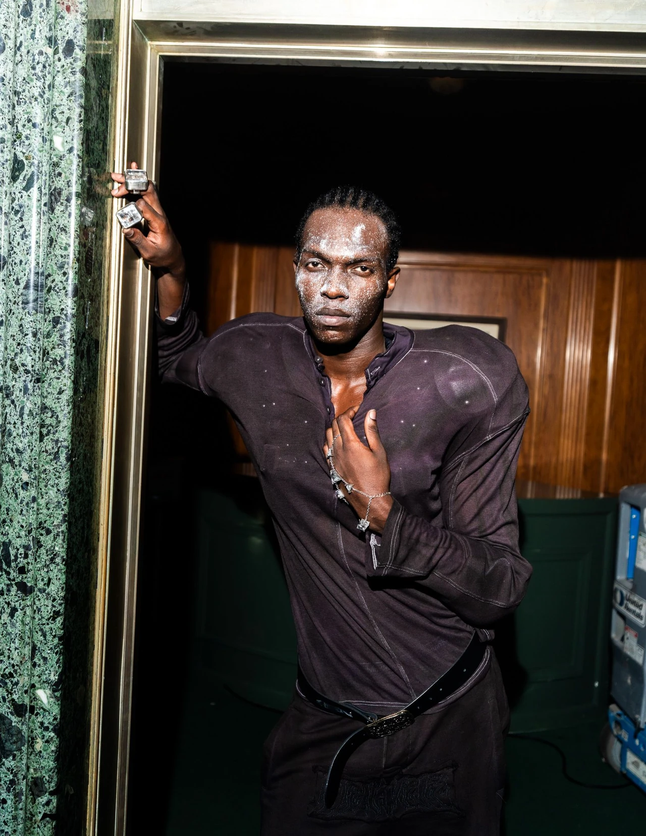 Model wearing a dark sheer top with sculpted shoulders and an open neckline in a green doorway.