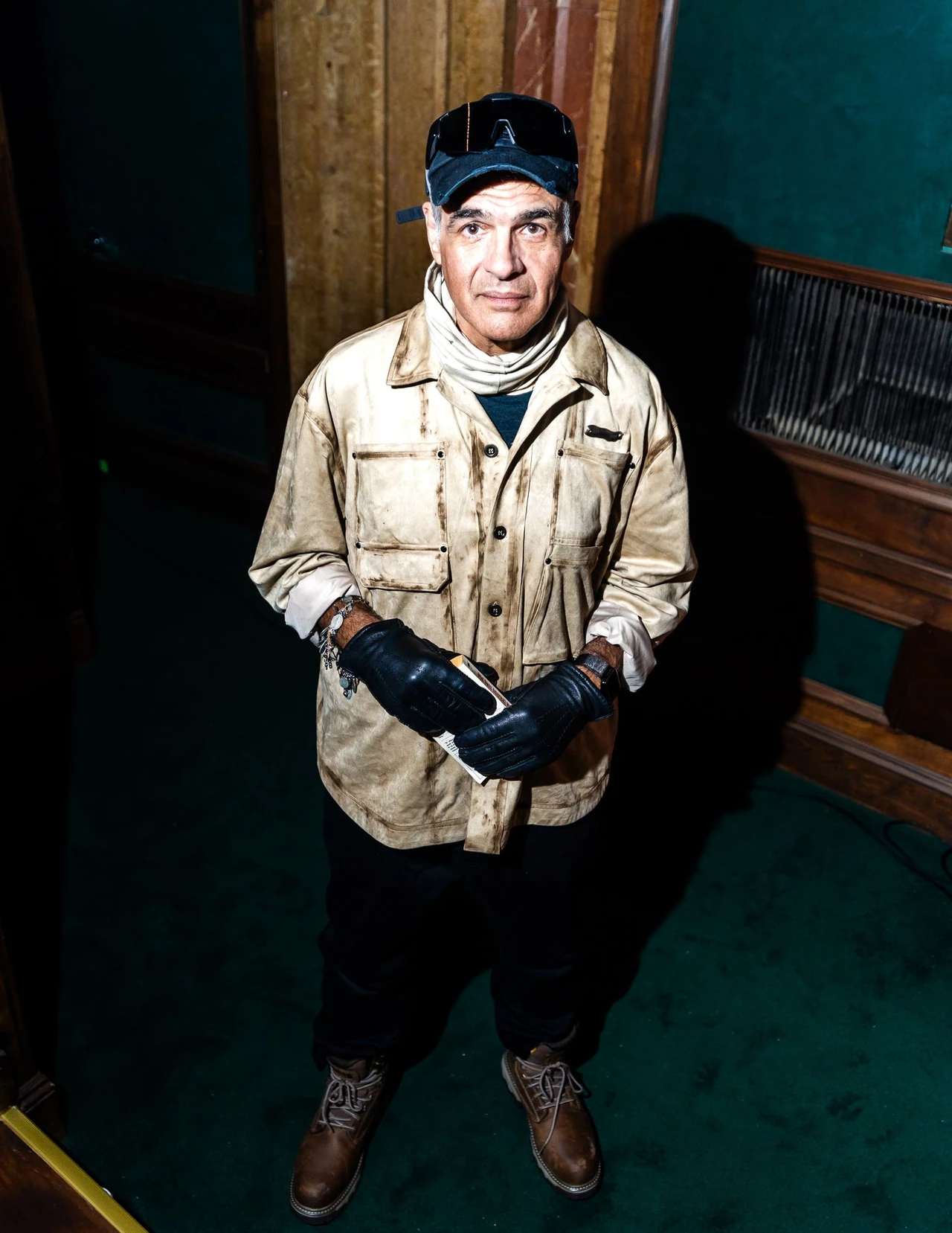 Model in a distressed tan jacket, cap and gloves standing against wood paneling.