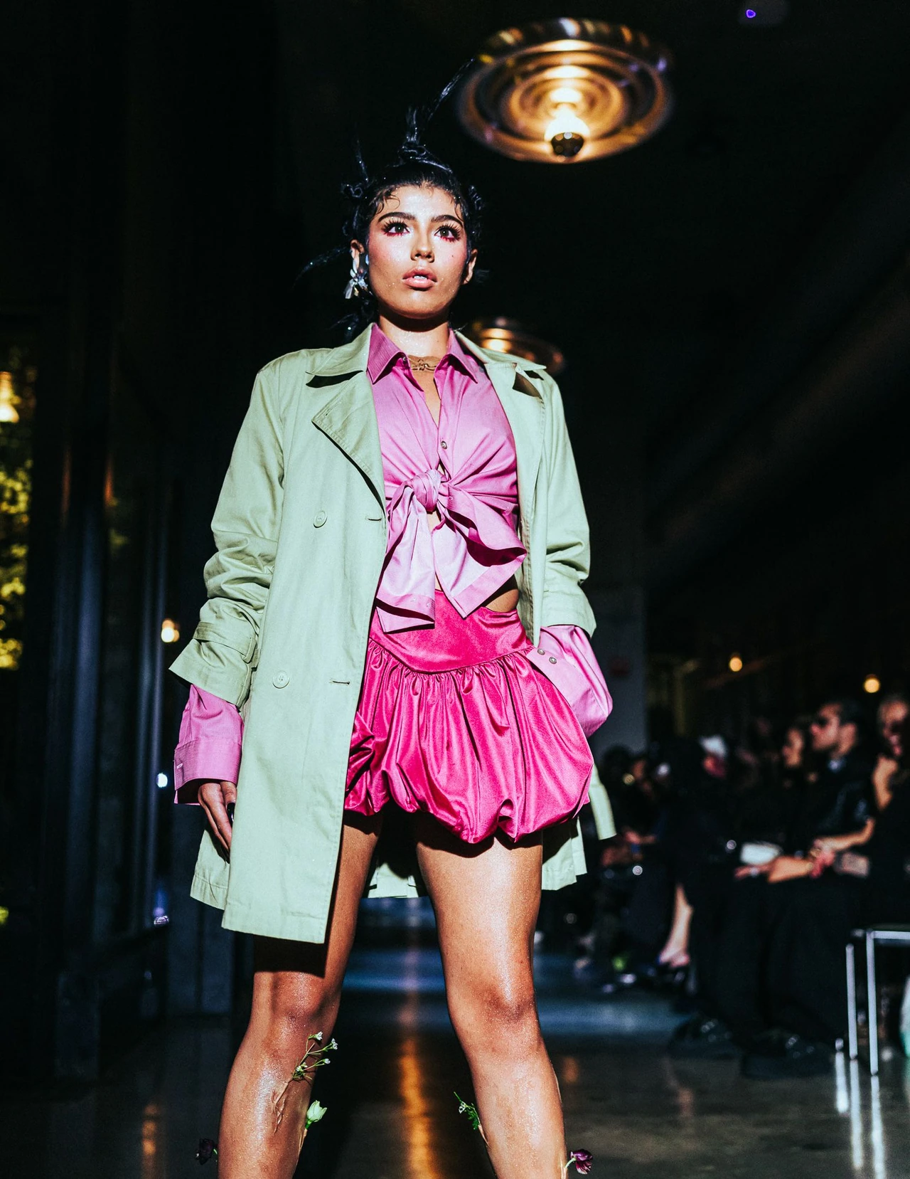 Model wearing a green blazer over a pink blouse and bubble shorts on a dark runway.