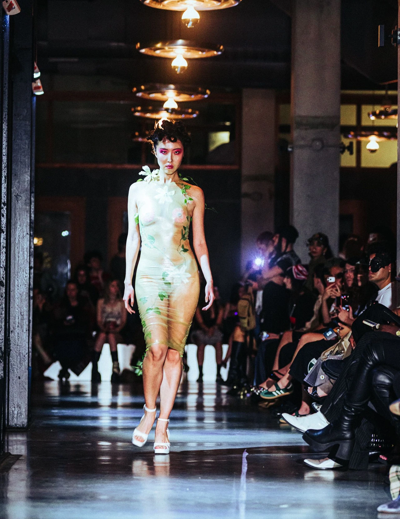 RIO runway model in a pale green sculptural outfit walking between seated guests beneath round ceiling lights.