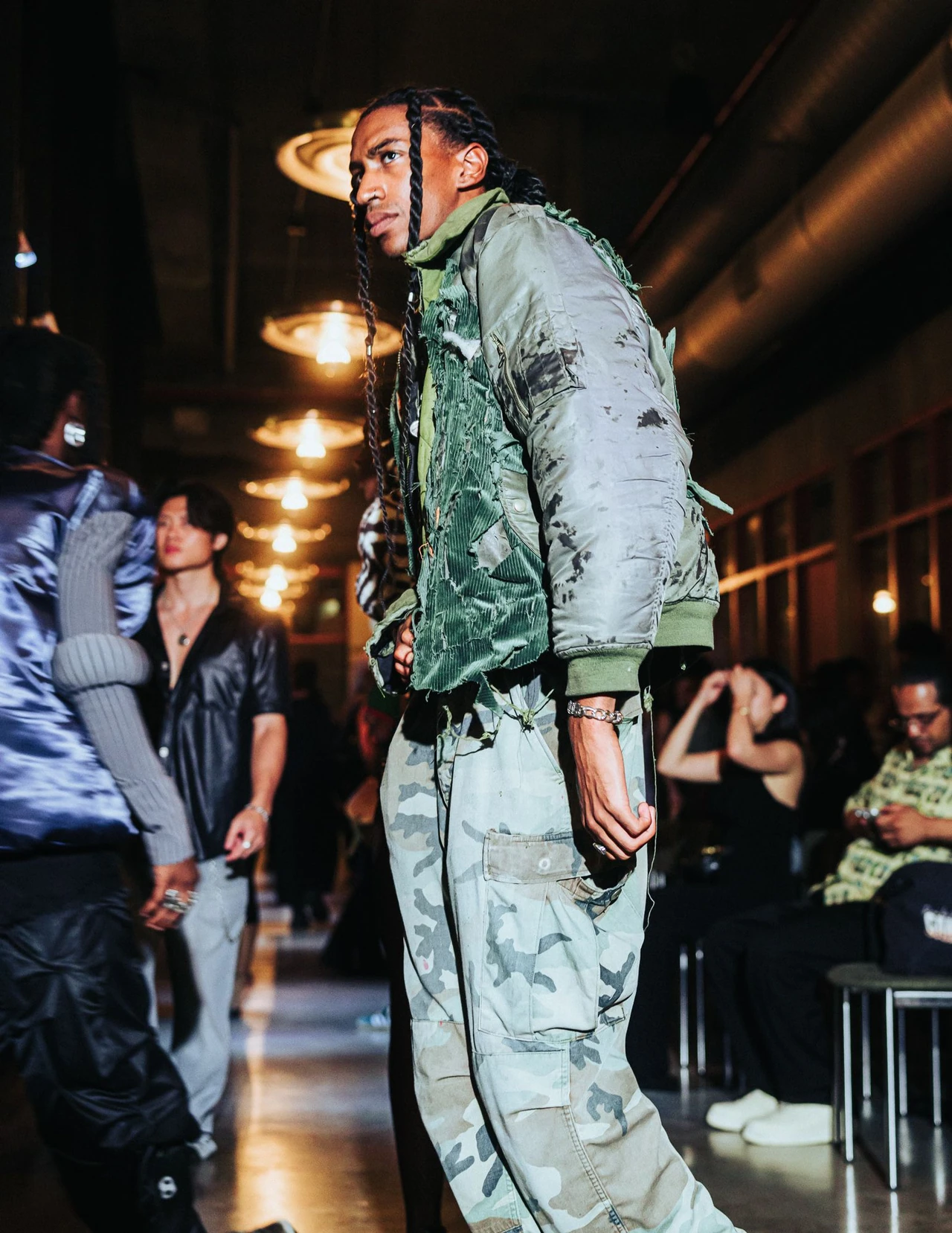 Model in a green bomber jacket and camouflage trousers turning toward the camera on the runway.