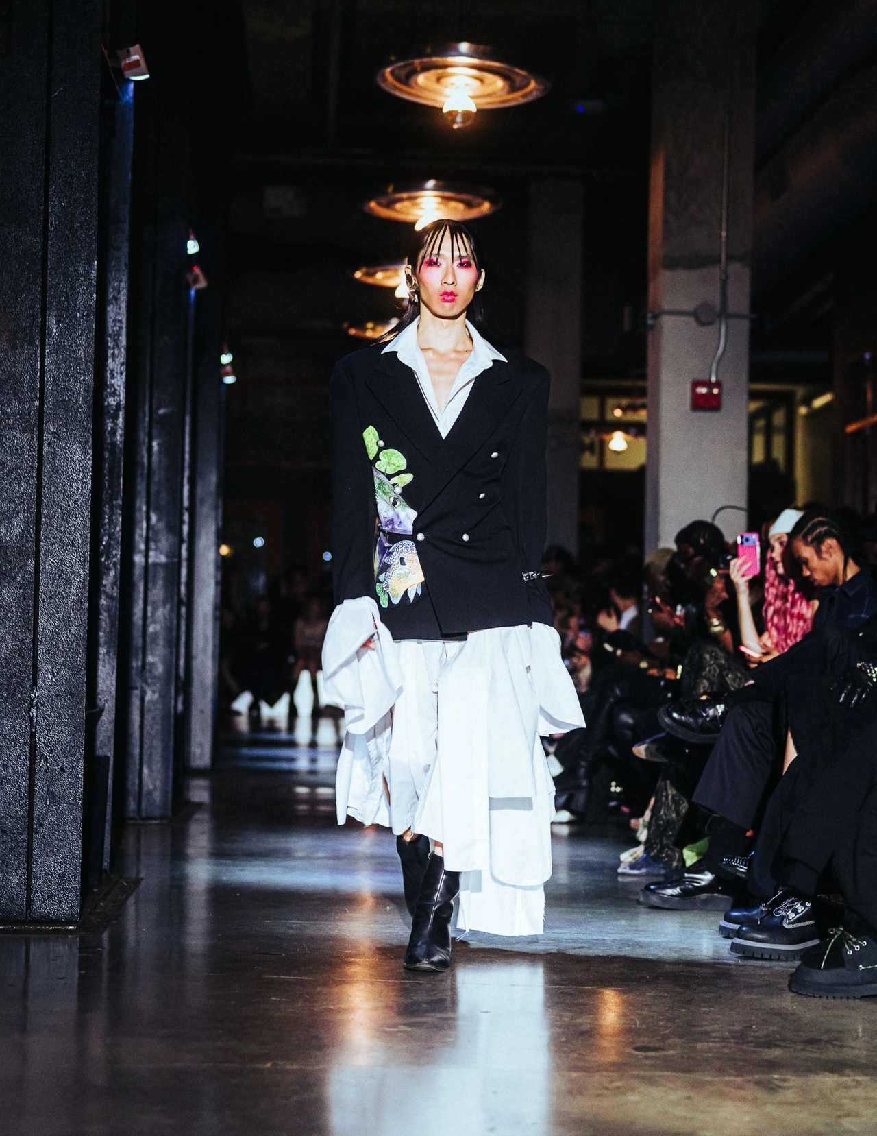 Model wearing a dark blazer with a white tie and layered white shorts on the runway.
