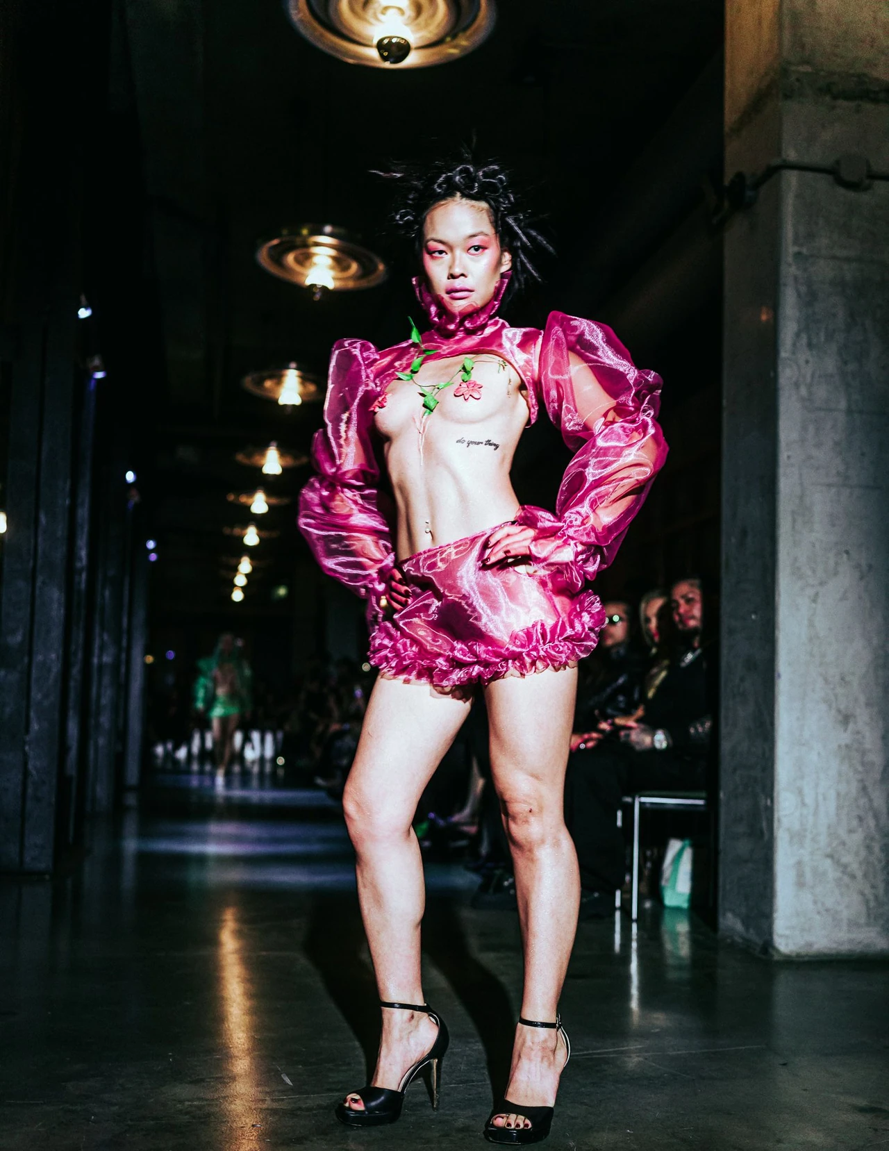 Model in a shiny pink cropped jacket and shorts walking beneath warm ceiling lights.