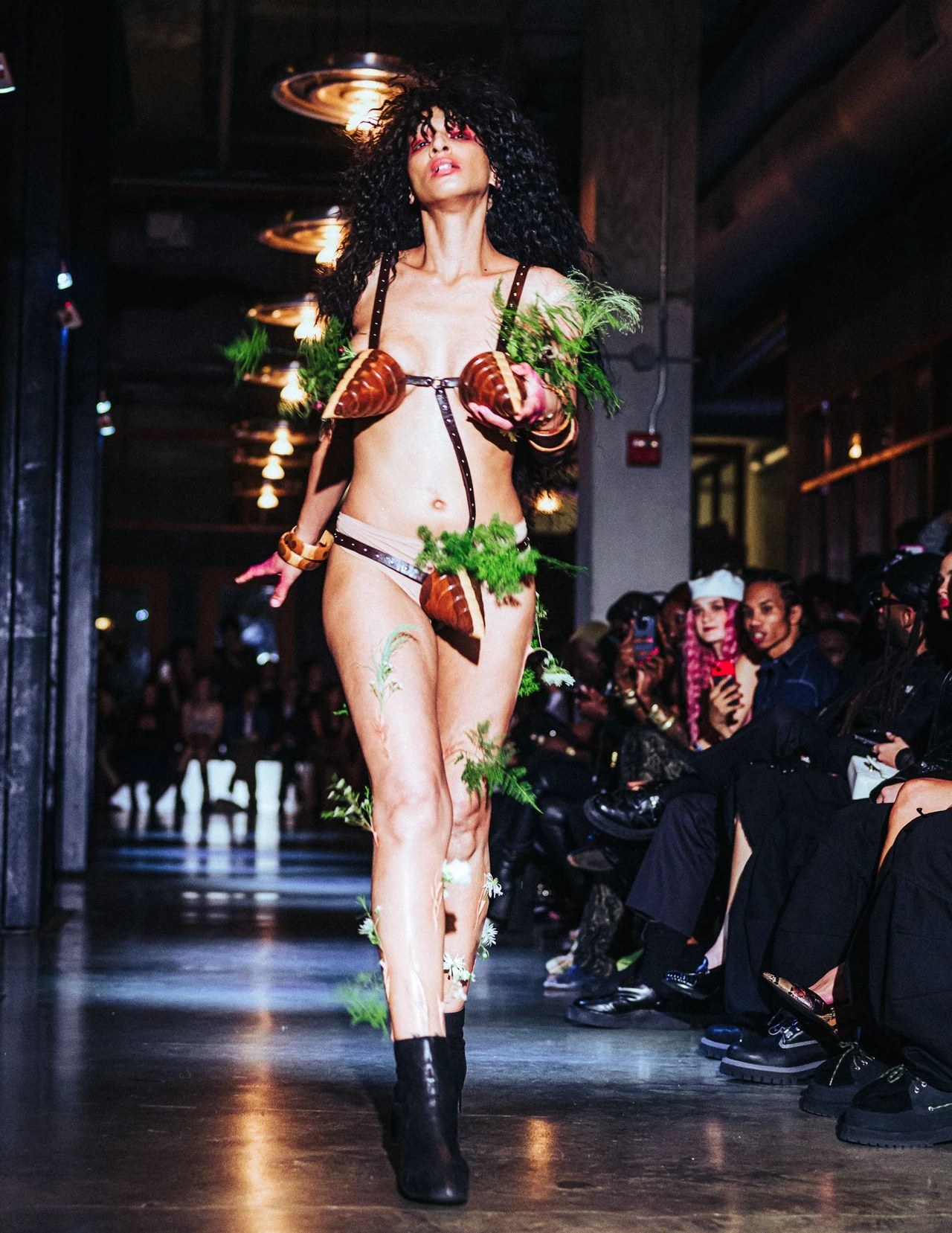 Model in a dark outfit with plant-like shoulder details walking between seated spectators.