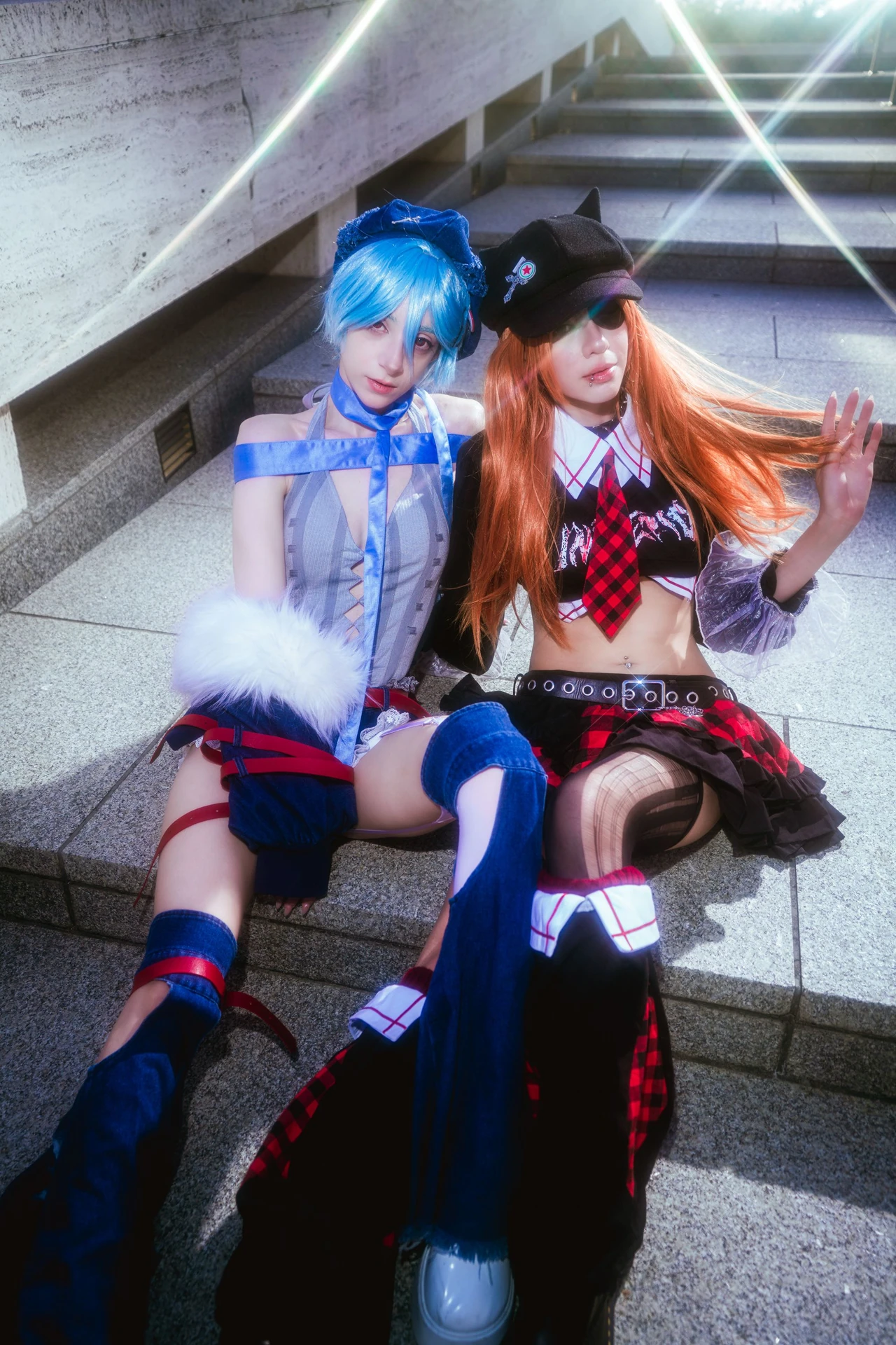 Rei and Asuka cosplay seated together on steps, with blue and orange wigs and matching dark caps.
