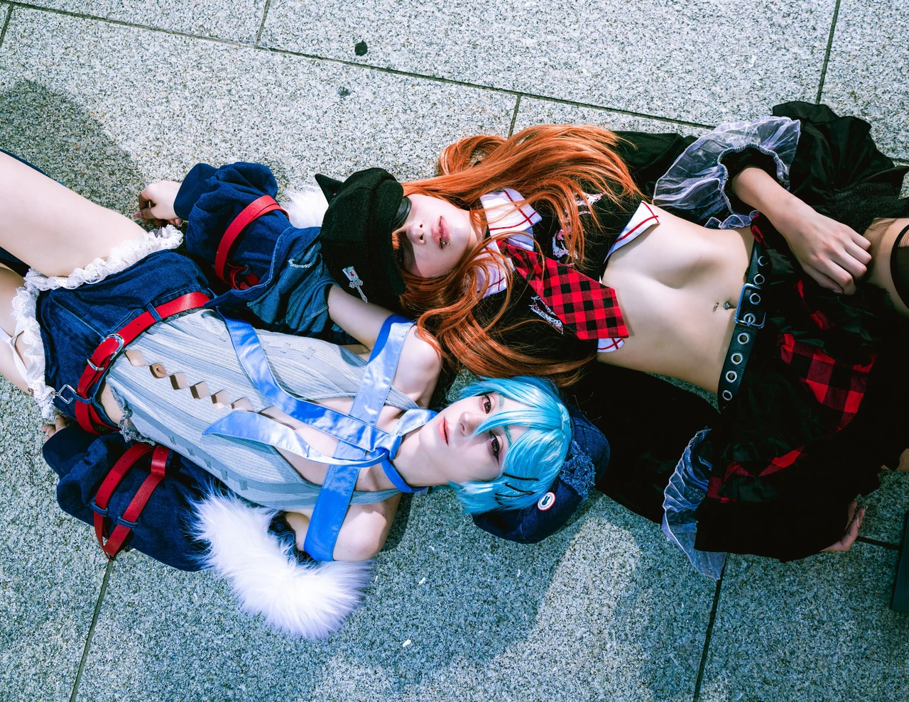 Overhead portrait of Rei and Asuka cosplay reclining together on textured concrete.