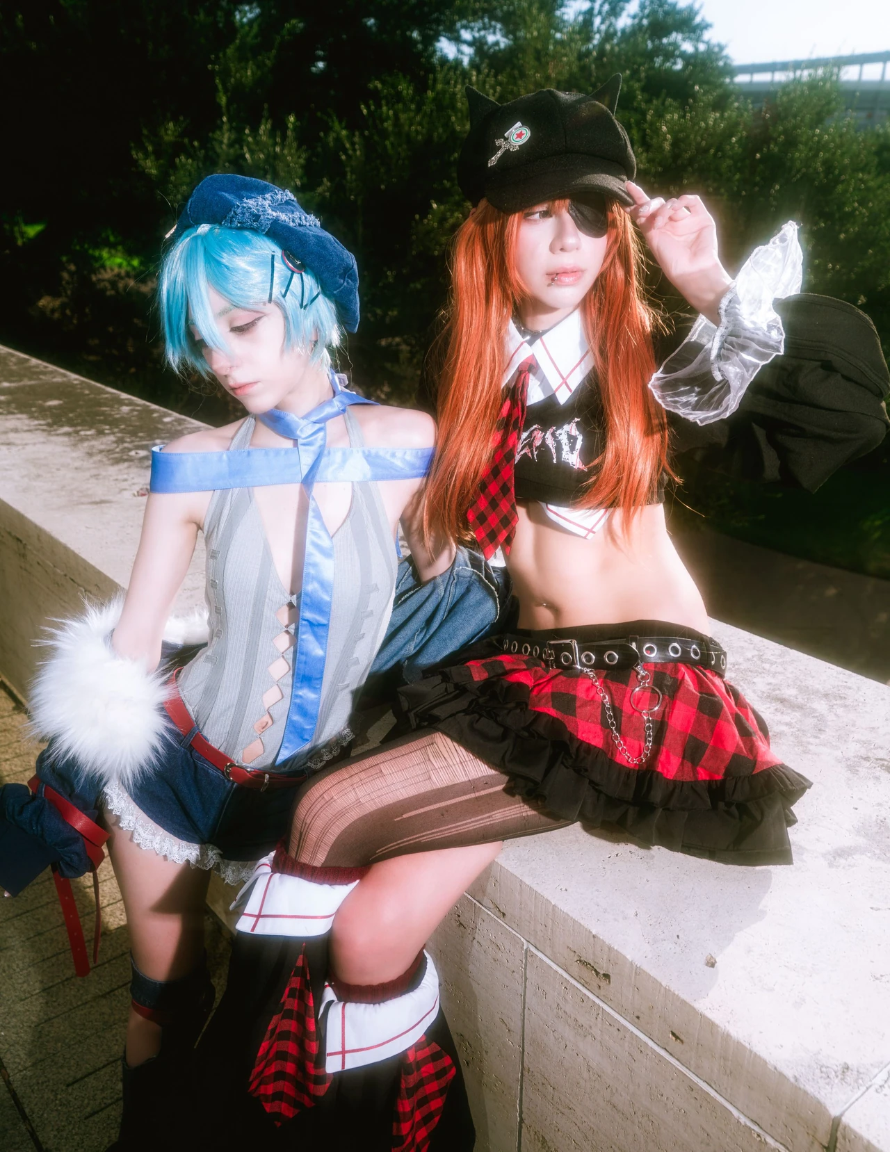 Orange-haired Asuka cosplay seated beside blue-haired Rei on a concrete ledge in daylight.