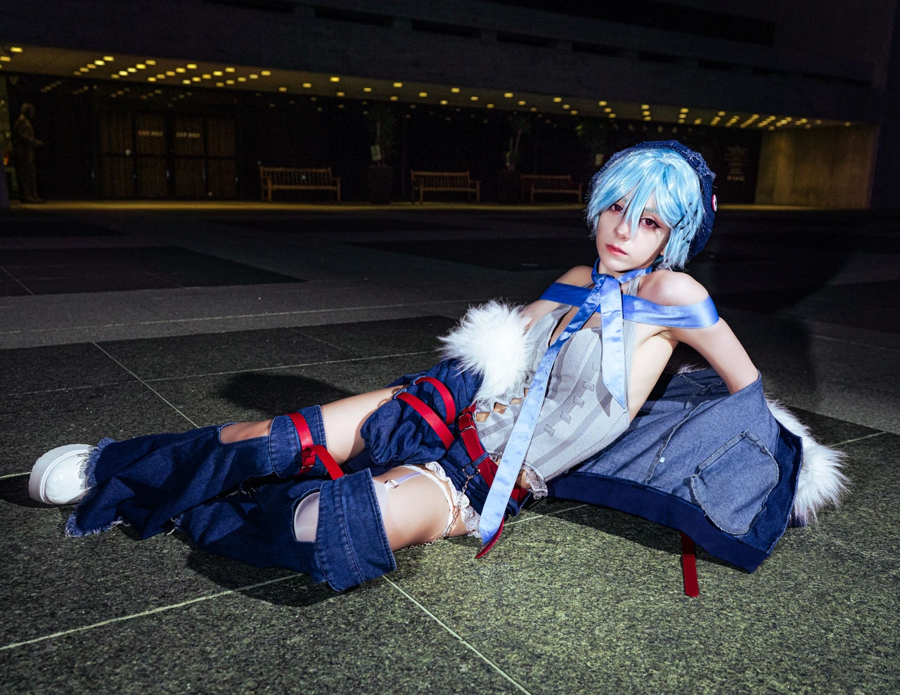 Rei cosplay seated on the ground in blue flared trousers against a dark evening background.