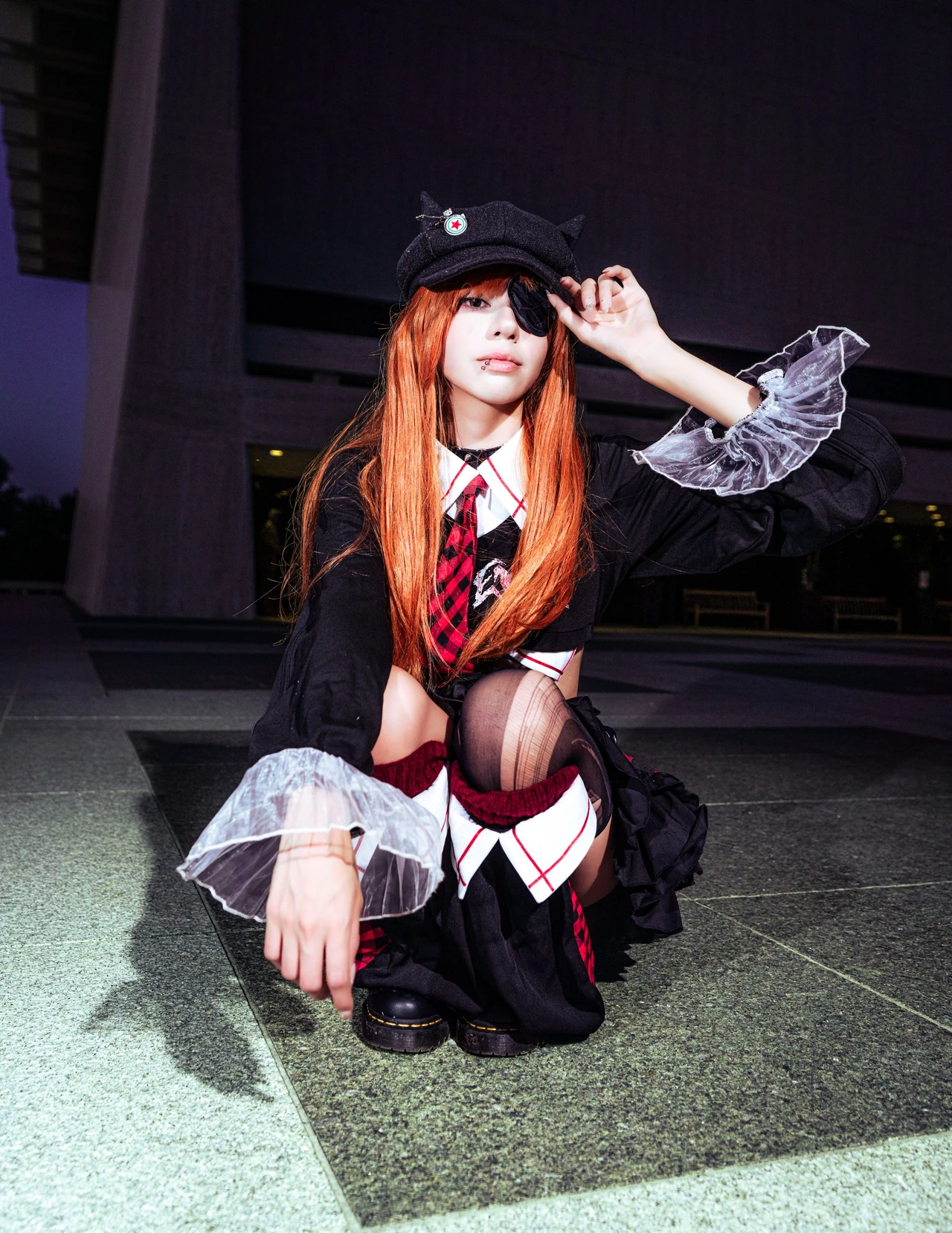 Asuka cosplay crouching in a red plaid skirt and dark cap with hands raised beside the head.