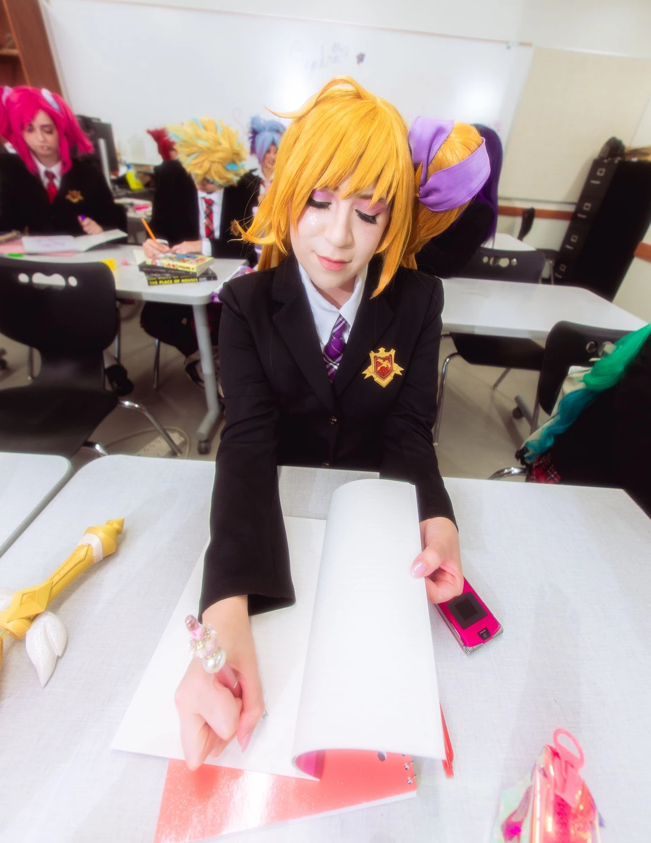 Cosplayer with golden hair and pointed ears holding a sheet of paper at a classroom desk.