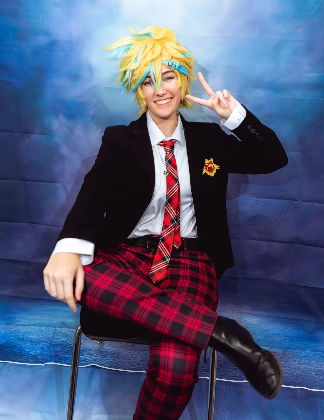 Yellow-haired cosplayer smiling and making a peace sign against a blue portrait backdrop.