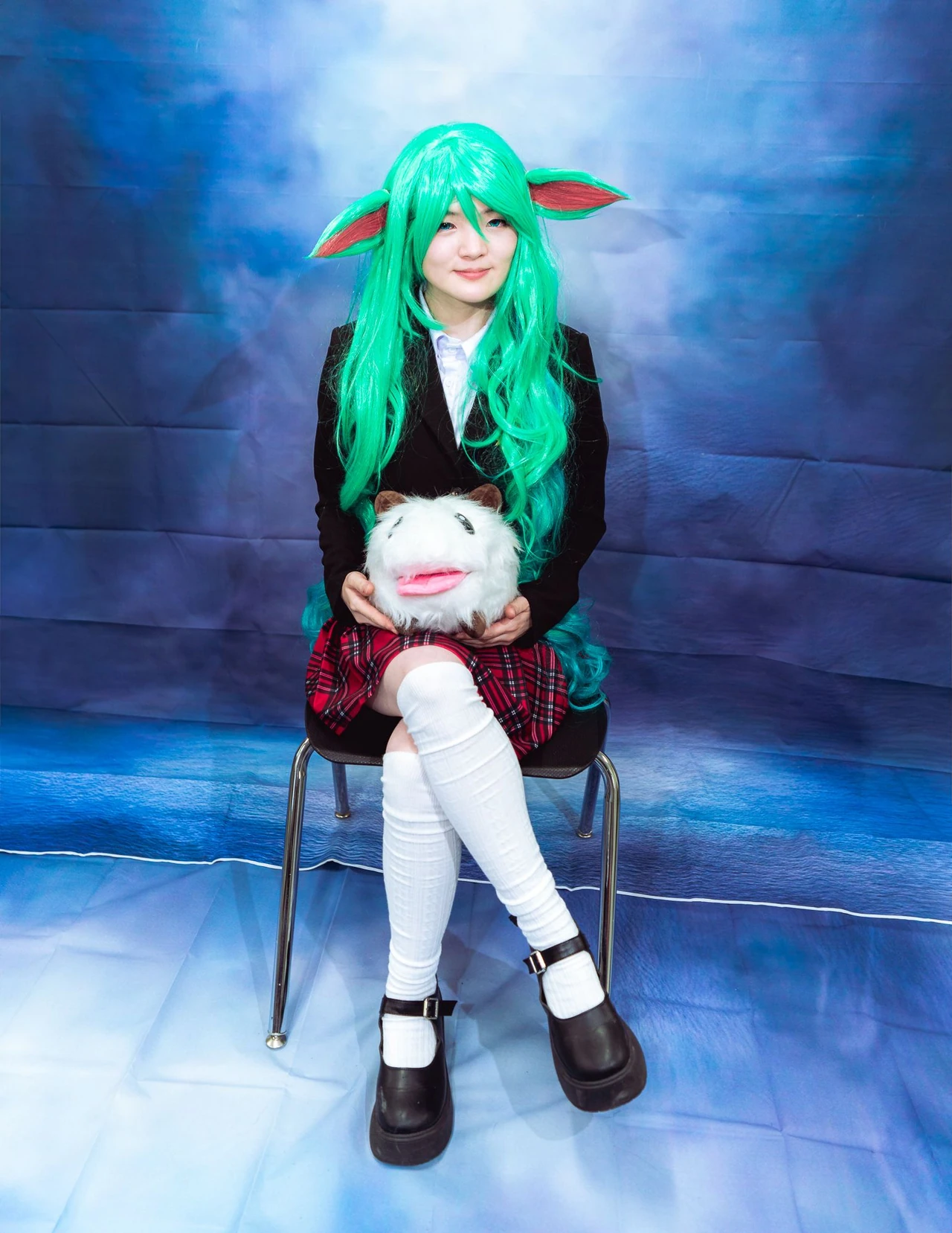 Green-haired cosplayer holding a plush toy while seated against a blue portrait backdrop.