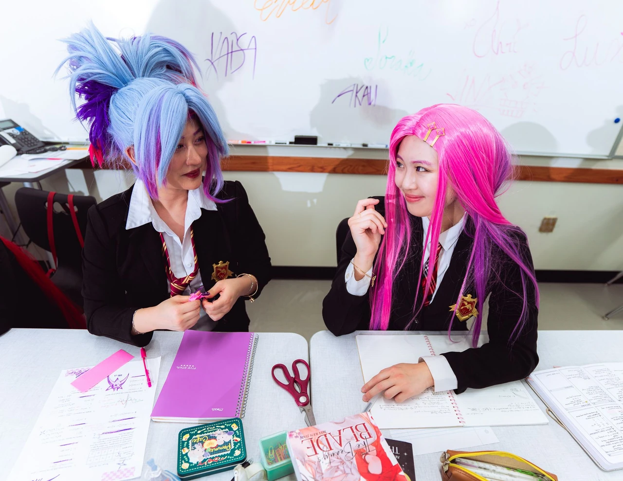 Two cosplayers with blue and pink wigs working together at a classroom desk.