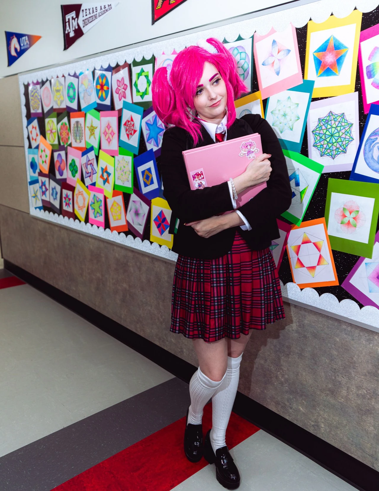 Pink-haired cosplayer holding a notebook beside a colorful school display and lockers.
