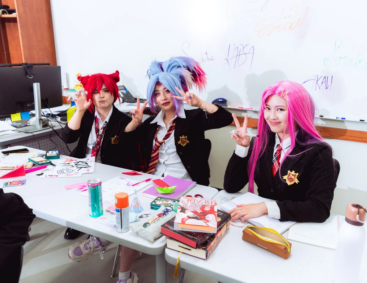 Three cosplayers in school uniforms holding snacks and posing beside classroom papers.