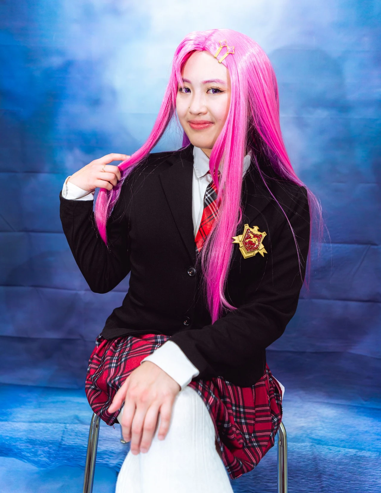 Pink-haired cosplayer smiling with a hand raised beside the face against a blue backdrop.