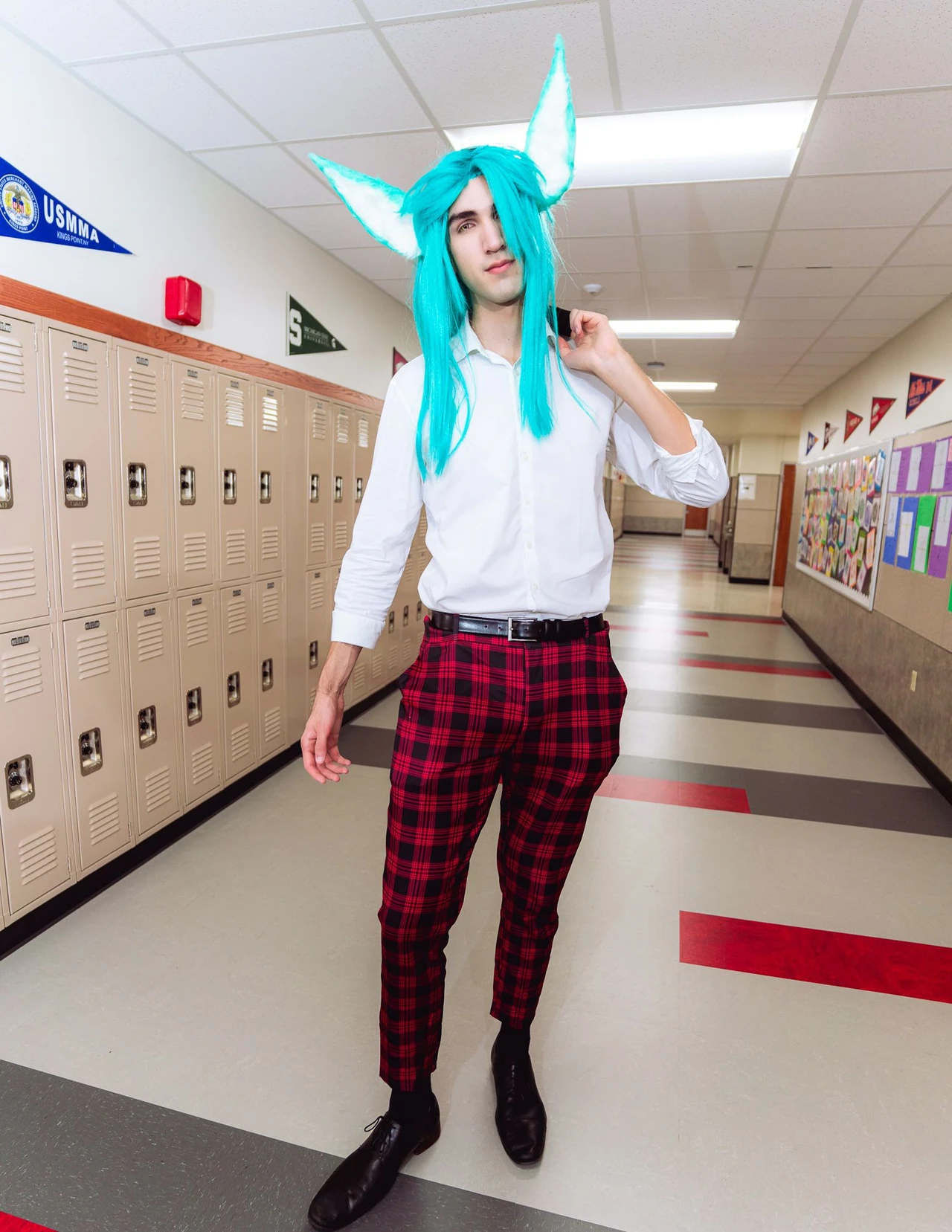 Turquoise-haired cosplayer in a white sleeveless shirt and plaid trousers standing in a school corridor.