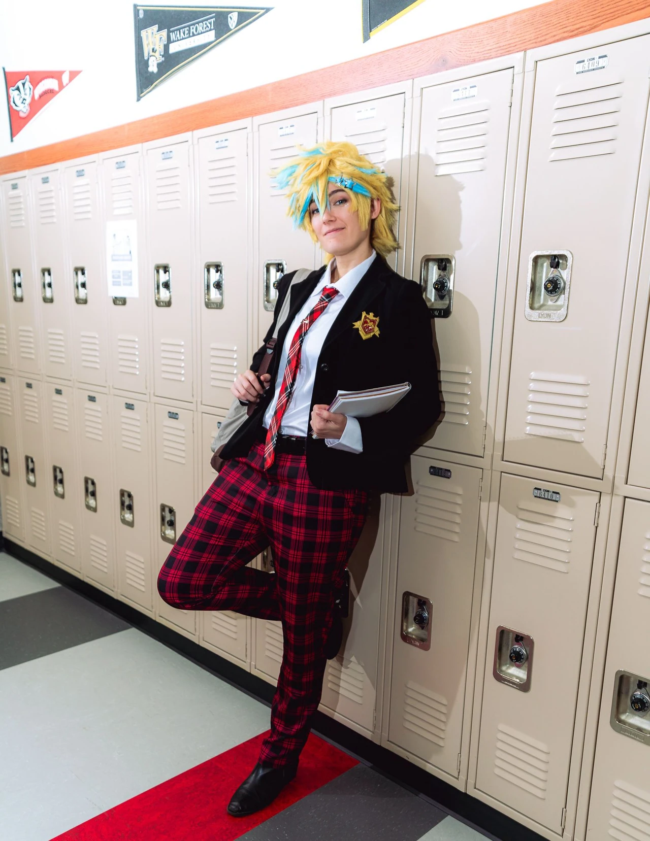Yellow-haired cosplayer in a black blazer and red plaid trousers standing beside school lockers.