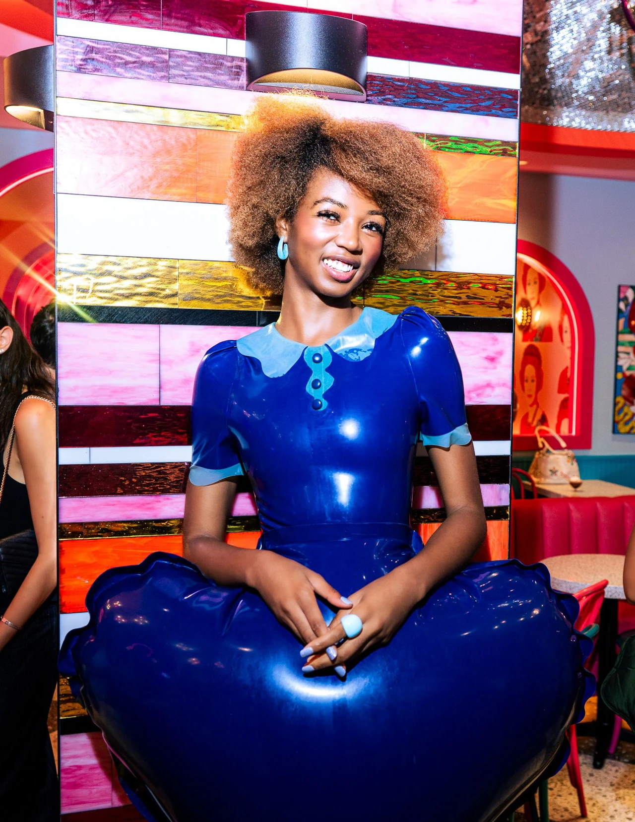 Model smiling in a glossy blue inflated dress against striped restaurant walls.