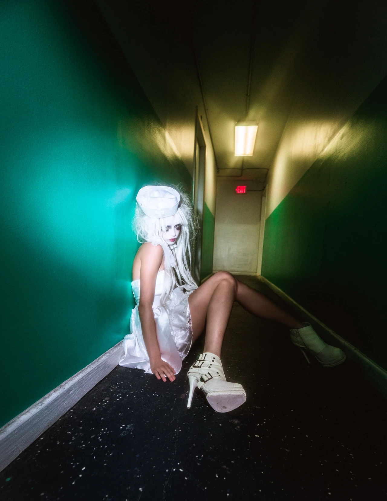 Model in white seated on the floor of a dark green corridor with platform boots extended.