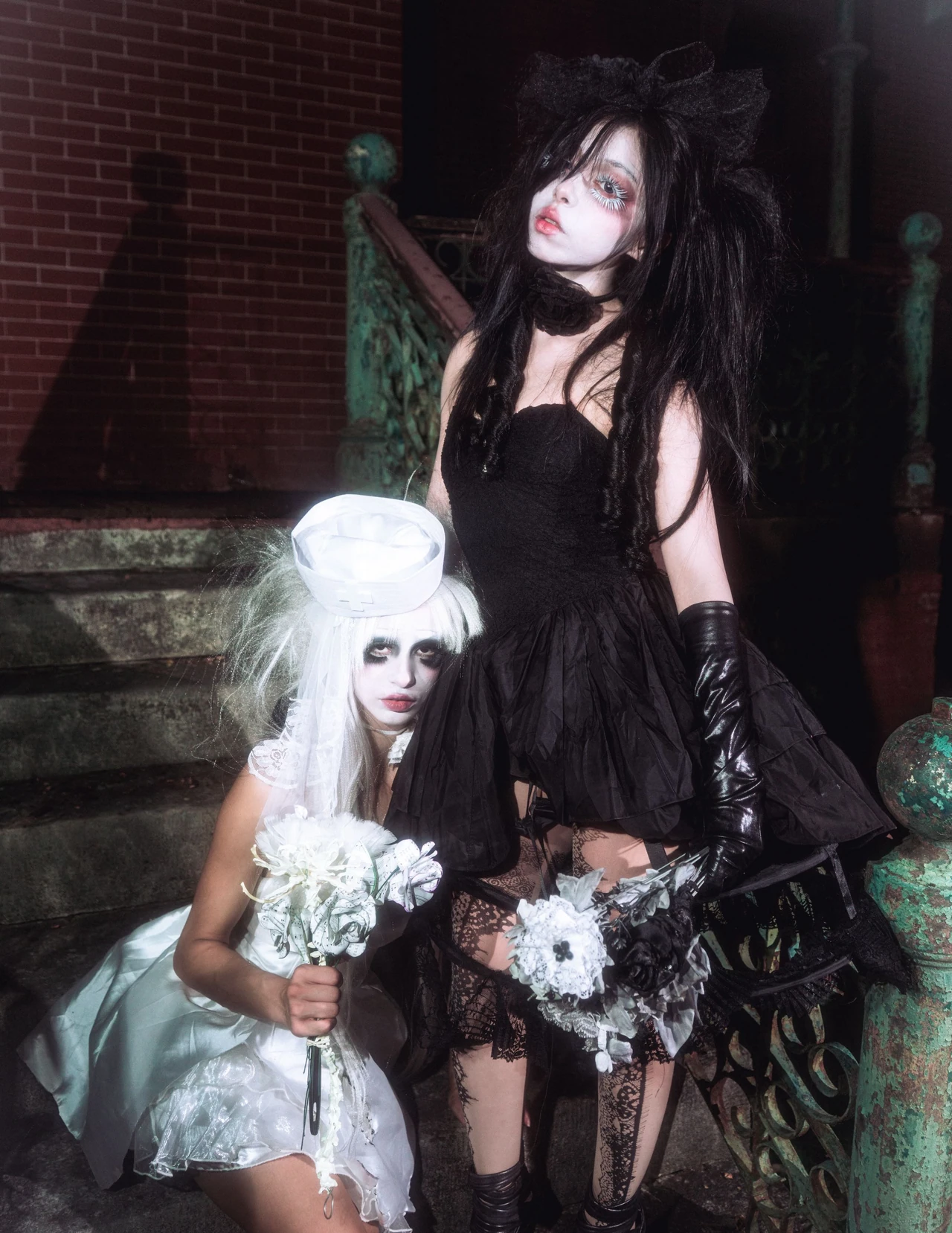 Two models in black and white outfits with pale makeup and flowers framed by a green railing.