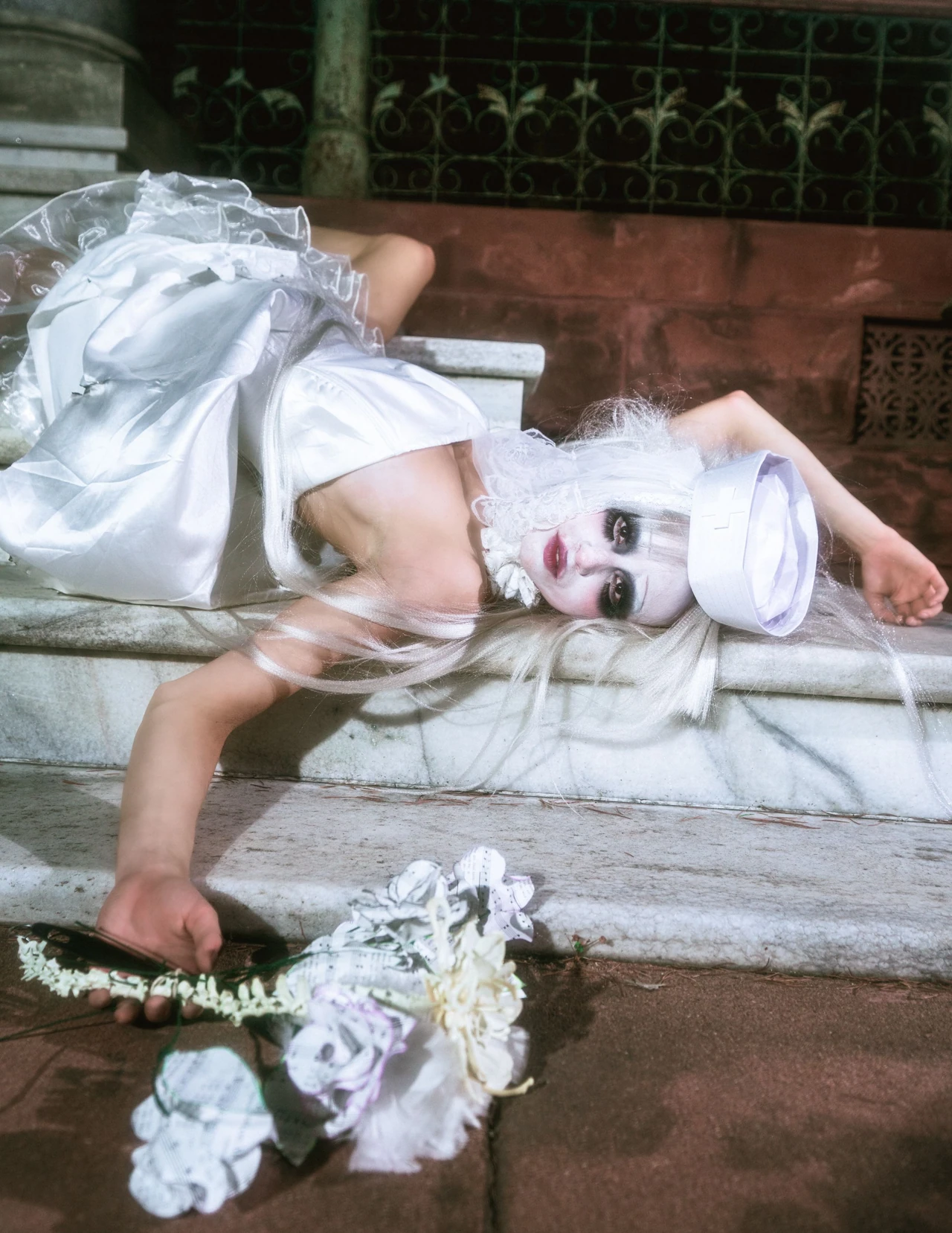 Model in white reclining on pale stone steps with a bouquet of flowers in the foreground.