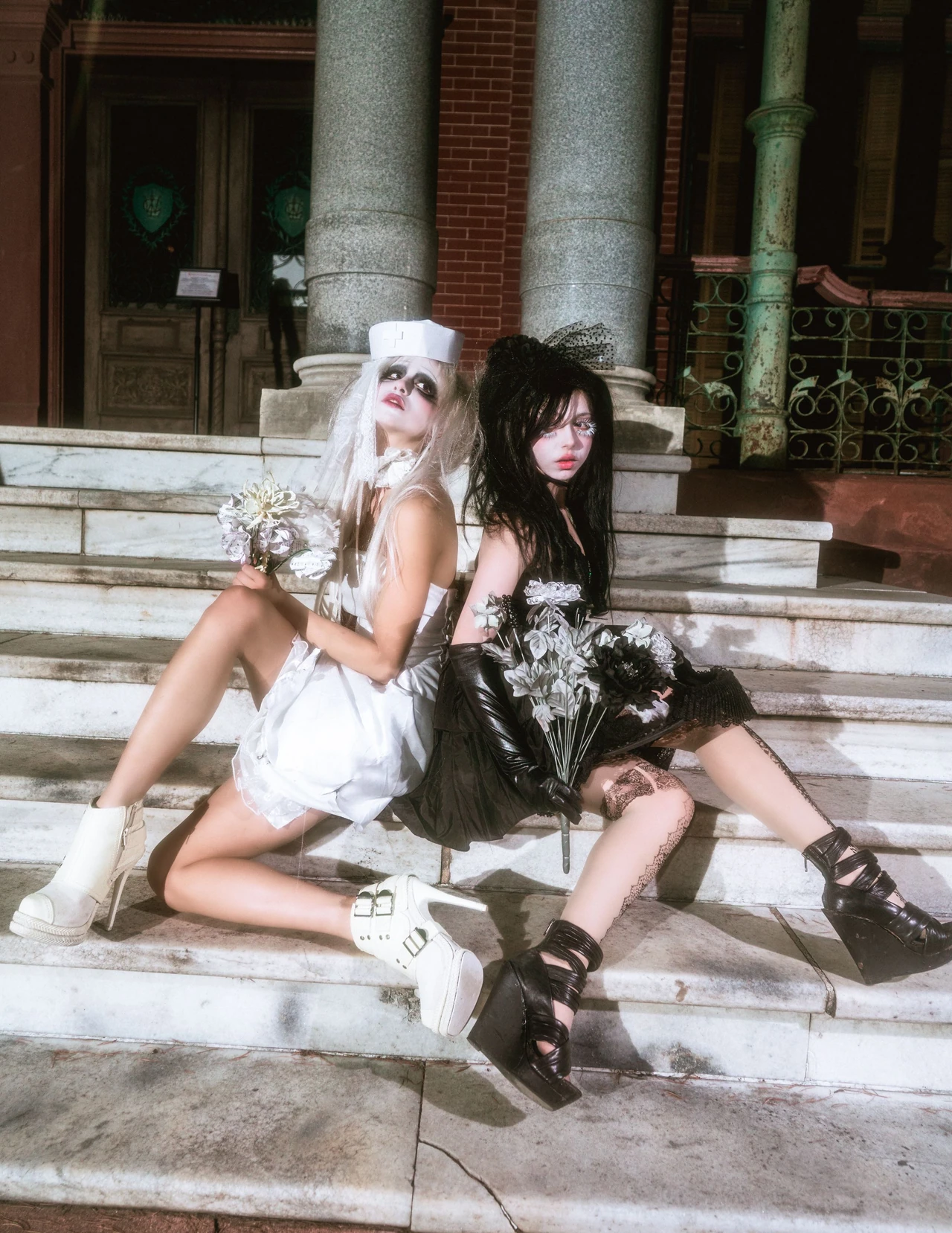 Models in white and black doll-inspired dresses seated back to back on stone steps.
