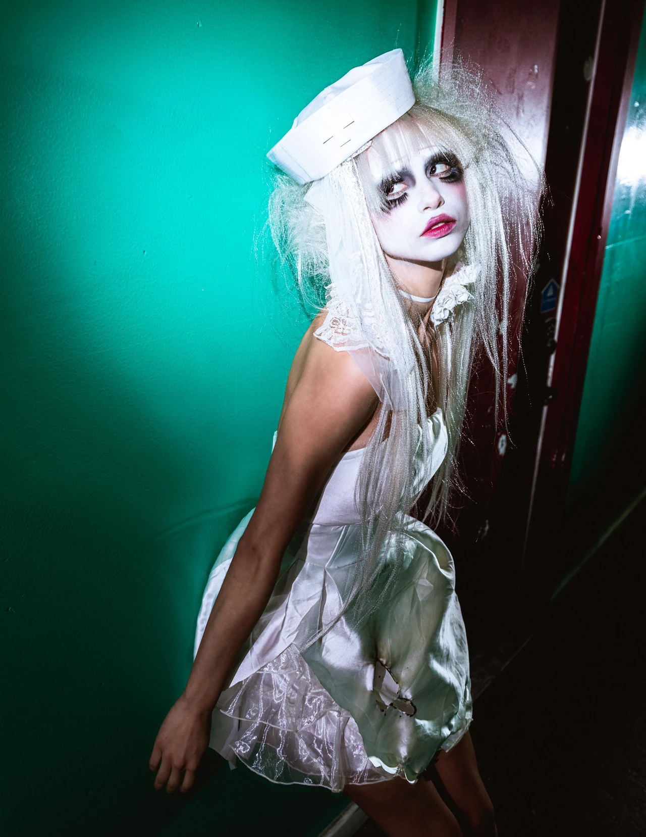 Model in a white dress and tiny hat looking up inside a narrow green corridor.