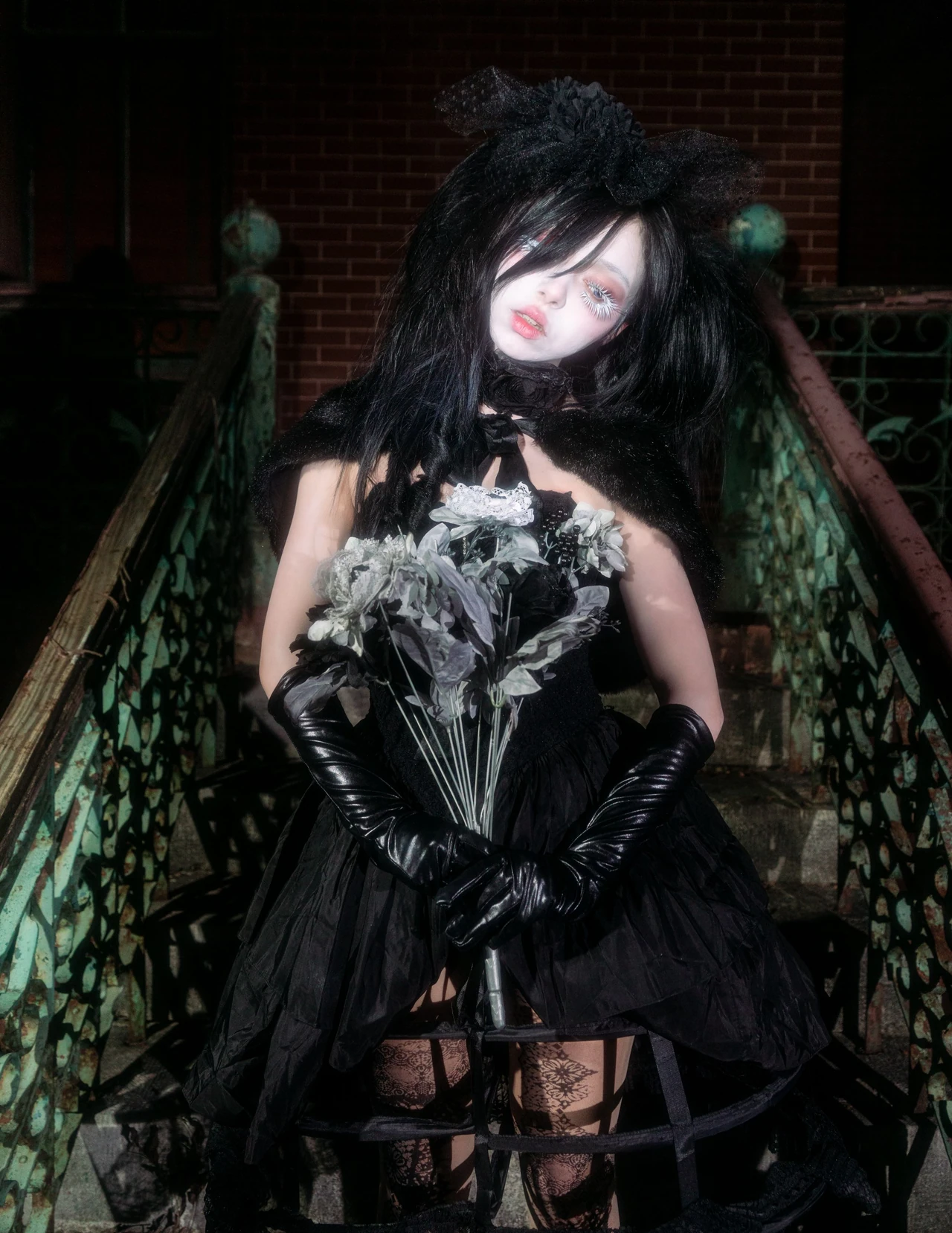 Model in a black lace dress and gloves holding flowers beside an ornate iron railing at night.
