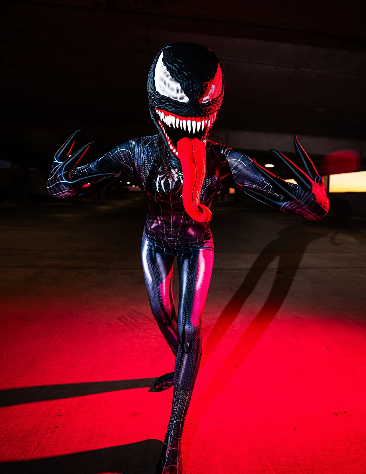 Venom cosplayer posing with raised claws and a long red tongue in a dark parking garage.