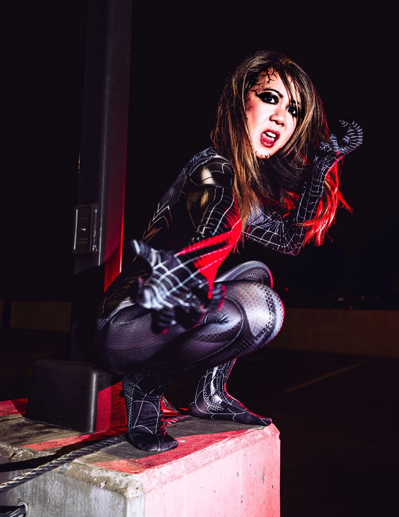 Unmasked cosplayer in a black Venom suit crouching on a concrete ledge with red light in the hair.